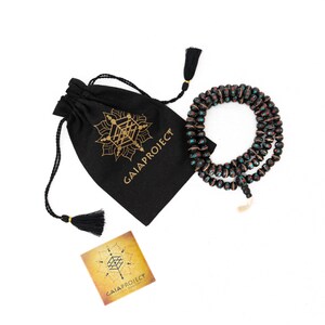Puede incluir: Una bolsa negra de cord&oacute;n con el texto "GAIA PROJECT" en letras doradas. La bolsa est&aacute; al lado de un collar de cuentas negras y verdes con acentos rojos. El collar est&aacute; sobre un fondo blanco.