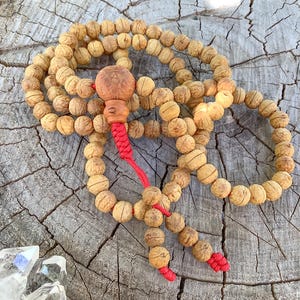 Puede incluir: Un collar de cuentas de oración con cuentas redondas de color marrón claro y una pieza central de madera tallada. Las cuentas están ensartadas en un cordón rojo, con una borla en el extremo. Las cuentas descansan sobre una superficie de madera envejecida y texturizada.