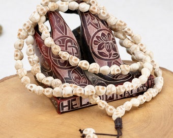May include: White skull bead necklace with a brown cord. The necklace is draped over a carved wooden stand with a floral design.