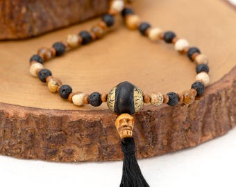 May include: A black and brown beaded necklace with a skull charm and a black tassel. The necklace is made with lava rock, jasper, and wood beads.