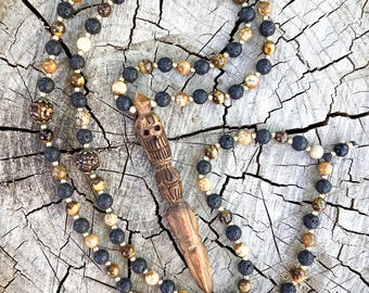May include: A handcrafted necklace featuring a carved wooden pendant and a beaded design. The beads are a mix of black, brown, and cream colors. The necklace is displayed on a weathered, textured wooden surface.