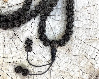 May include: A dark brown beaded necklace with a textured surface. The beads are strung together with a black cord, and the necklace is displayed on a light wood surface.