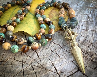May include: A beaded necklace with a gold-colored pendant. The necklace features a variety of earth-toned beads in shades of brown, tan, and green. The pendant is a detailed, ornate design. The necklace is displayed on a rustic wooden surface.