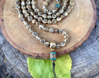 May include: A cream-colored mala bead necklace with black spiral designs. The necklace features turquoise accents and a decorative pendant with a black tassel. The beads are strung together, creating a spiritual or meditation accessory.