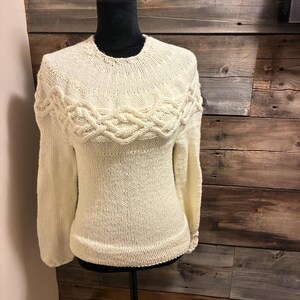 May include: Cream-coloured knitted sweater with a round neckline and long sleeves. The yoke features a decorative cable knit design. The sweater is displayed on a black mannequin against a wooden plank background.