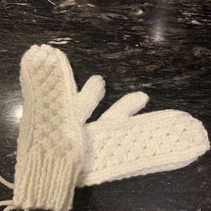 May include: A pair of white knitted mittens. The mittens are made of a thick, textured yarn with a diamond pattern on the back of the hand. The cuffs are ribbed, and a white string is attached to the wrist.