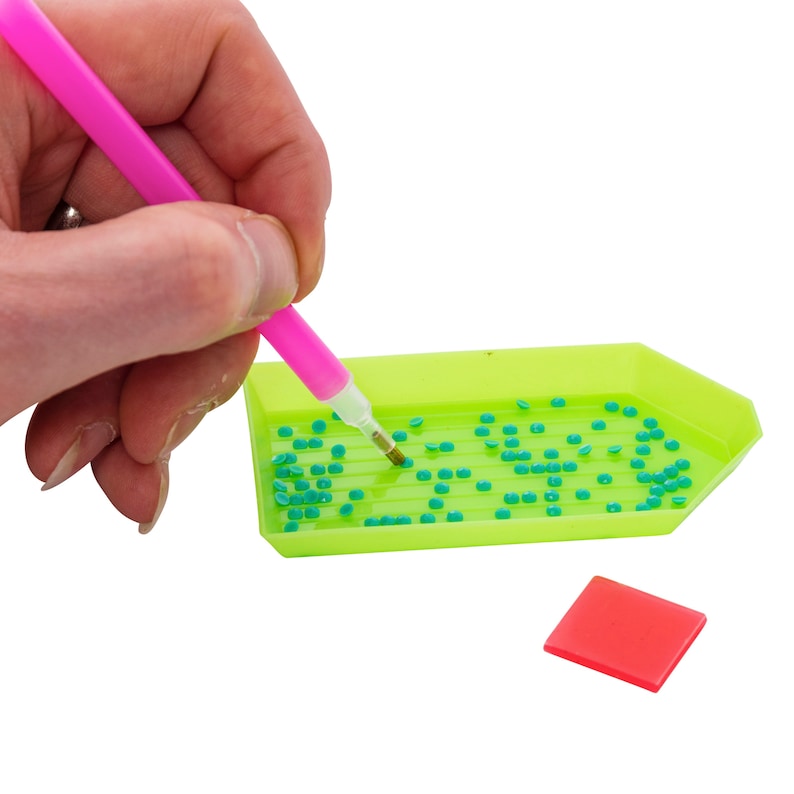 Puede incluir: Un bol&iacute;grafo rosa brillante para pintar con diamantes se utiliza para recoger peque&ntilde;as cuentas de diamantes de color verde azulado de una bandeja verde ne&oacute;n. Tambi&eacute;n se ve un peque&ntilde;o cuadrado rojo. La imagen est&aacute; sobre un fondo blanco.