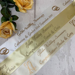May include: A selection of satin ribbons in white and gold, printed with gold text and graphics for golden wedding anniversaries. The ribbons feature phrases like "Golden Wedding Anniversary" and names. The background includes a pair of yellow roses.