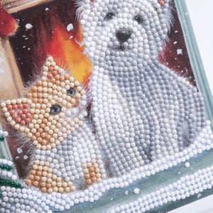 Puede incluir: Pintura de diamantes de un gatito y un cachorro, con una acogedora escena de chimenea en el fondo. El gatito es naranja y blanco, y el cachorro es gris y blanco. La imagen est&aacute; enmarcada con cuentas blancas.