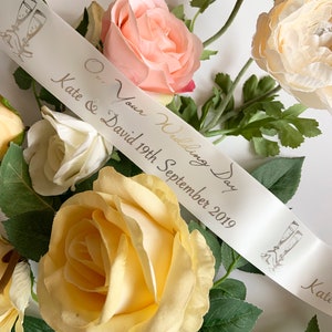 May include: Ivory satin ribbon with silver text reading "Kate & David 19th September 2019" and "On Your Wedding Day". Features illustrations of champagne flutes. Surrounded by peach, cream, and yellow roses.