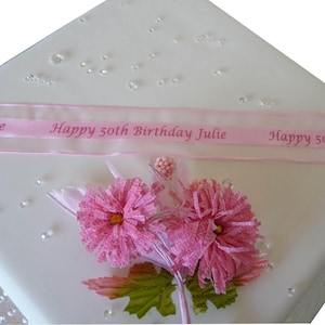 Puede incluir: Un pastel blanco cuadrado decorado con una cinta rosa que dice "Happy 50th Birthday Julie". El pastel está adornado con flores de tela rosa, hojas verdes y adornos brillantes y transparentes.