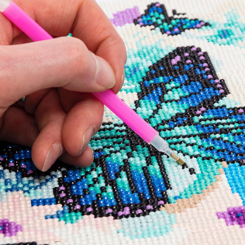 Puede incluir: Primer plano de una pintura de diamantes en progreso, con un dise&ntilde;o de mariposa en tonos azules, turquesas, negros y morados. Se utiliza un l&aacute;piz rosa para colocar peque&ntilde;as gemas en el lienzo. El proyecto de manualidades est&aacute; sobre un fondo blanco.
