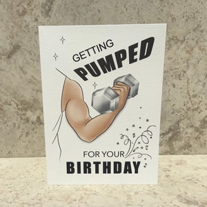 May include: A white card with a black and white illustration of a person lifting a dumbbell. The text on the card reads "Getting Pumped for Your Birthday".