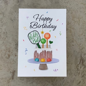 May include: A white card with the text "Happy Birthday" in black cursive lettering. A brown and white cake with white frosting and a pickleball paddle on top is on a purple cake stand. The cake is decorated with colorful pickleballs.