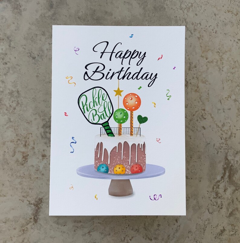 May include: A white card with the text "Happy Birthday" in black cursive lettering. A brown and white cake with white frosting and a pickleball paddle on top is on a purple cake stand. The cake is decorated with colorful pickleballs.