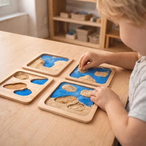 Può includere: Quattro vassoi in legno in stile Montessori con sabbia e riempimento blu, che simulano terra e acqua. Le mani di un bambino interagiscono con la sabbia, disponendo le forme. I vassoi sono su un tavolo di legno chiaro, con una mensola di legno sullo sfondo.