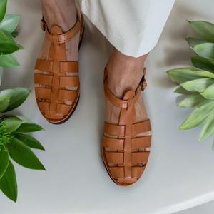 May include: Tan leather sandals with a woven design and an ankle strap. The sandals have a closed toe and a flat sole.