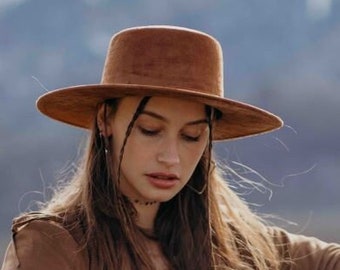 May include: A woman wearing a brown suede jacket with fringe, a brown felt hat, blue jeans, and brown boots. She is sitting on a tree stump.