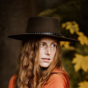 May include: A woman with long, wavy red hair wears a rust-colored wrap dress and a wide-brimmed black hat. She is standing in front of a background of yellow and green leaves.