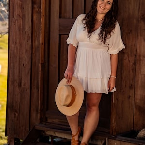 May include: A woman in a white dress stands on wooden steps, holding a straw hat. She is smiling and looking at the camera. The background is a wooden wall.