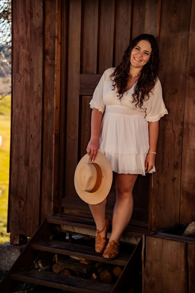 May include: A woman in a white dress stands on wooden steps, holding a straw hat. She is smiling and looking at the camera. The background is a wooden wall.