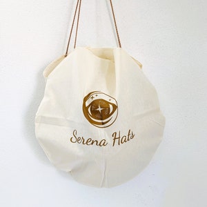 Peut inclure: Un sac en toile blanc avec des sangles brunes et un logo dor&eacute; qui dit "Serena Hats" avec un design d'&oelig;il stylis&eacute;.