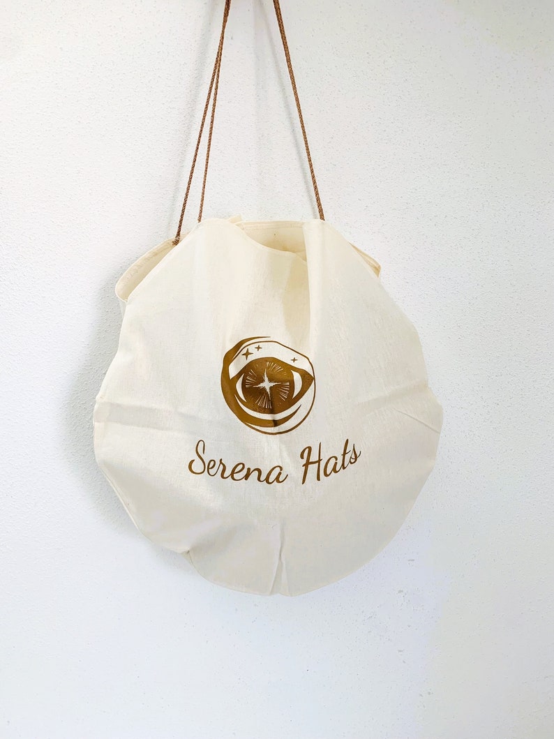Peut inclure: Un sac en toile blanc avec des sangles brunes et un logo dor&eacute; qui dit "Serena Hats" avec un design d'&oelig;il stylis&eacute;.