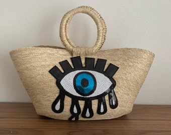Puede incluir: Un bolso de paja con un gran ojo azul con pestañas y lentejuelas en forma de lágrima. El bolso tiene un asa tejida.