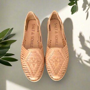 K&ouml;nnte beinhalten: Ein Paar braune Ledersandalen mit gewebten Details. Die Sandalen haben ein Slip-On-Design und der Markenname "SALT & SOHE" ist auf der Innensohle aufgedruckt.