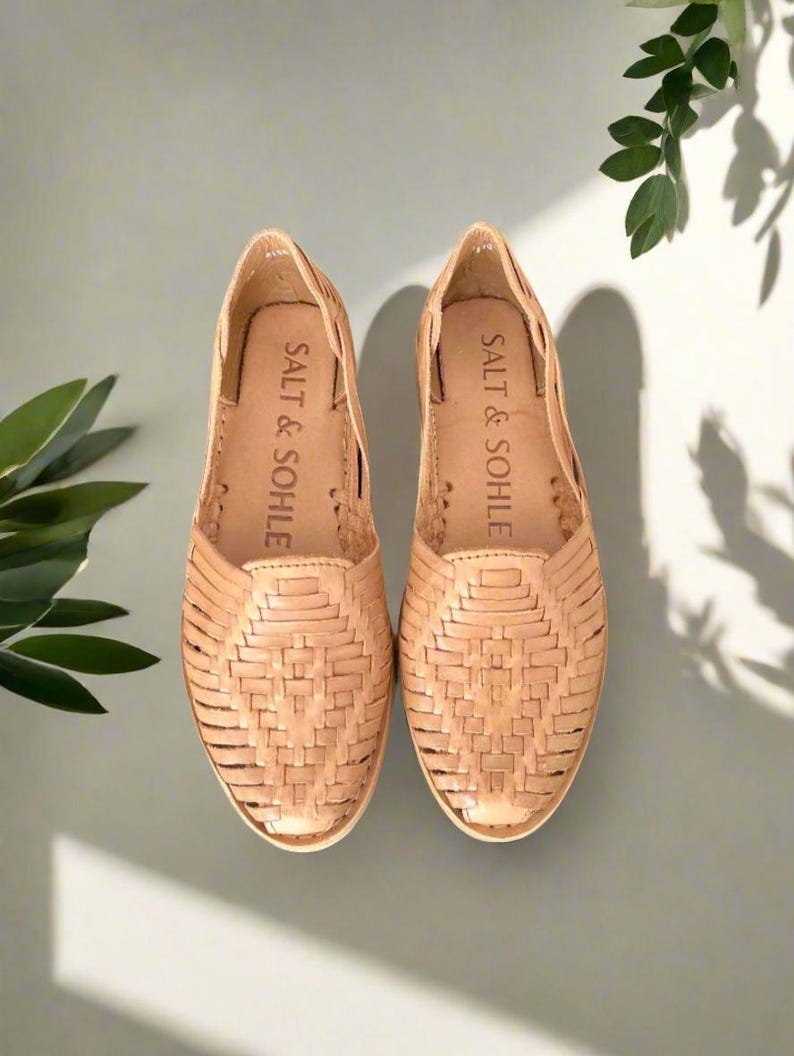 K&ouml;nnte beinhalten: Ein Paar braune Ledersandalen mit gewebten Details. Die Sandalen haben ein Slip-On-Design und der Markenname "SALT & SOHE" ist auf der Innensohle aufgedruckt.