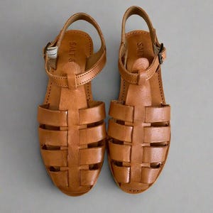 May include: A pair of brown leather sandals with a woven design. The sandals have a buckle closure and the brand name "Salt &" is visible on the strap.