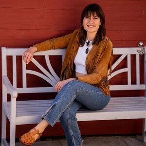 May include: A woman wearing a brown suede fringe jacket, white crop top, blue jeans, and brown sandals sits on a white wooden bench in front of a red wooden wall.