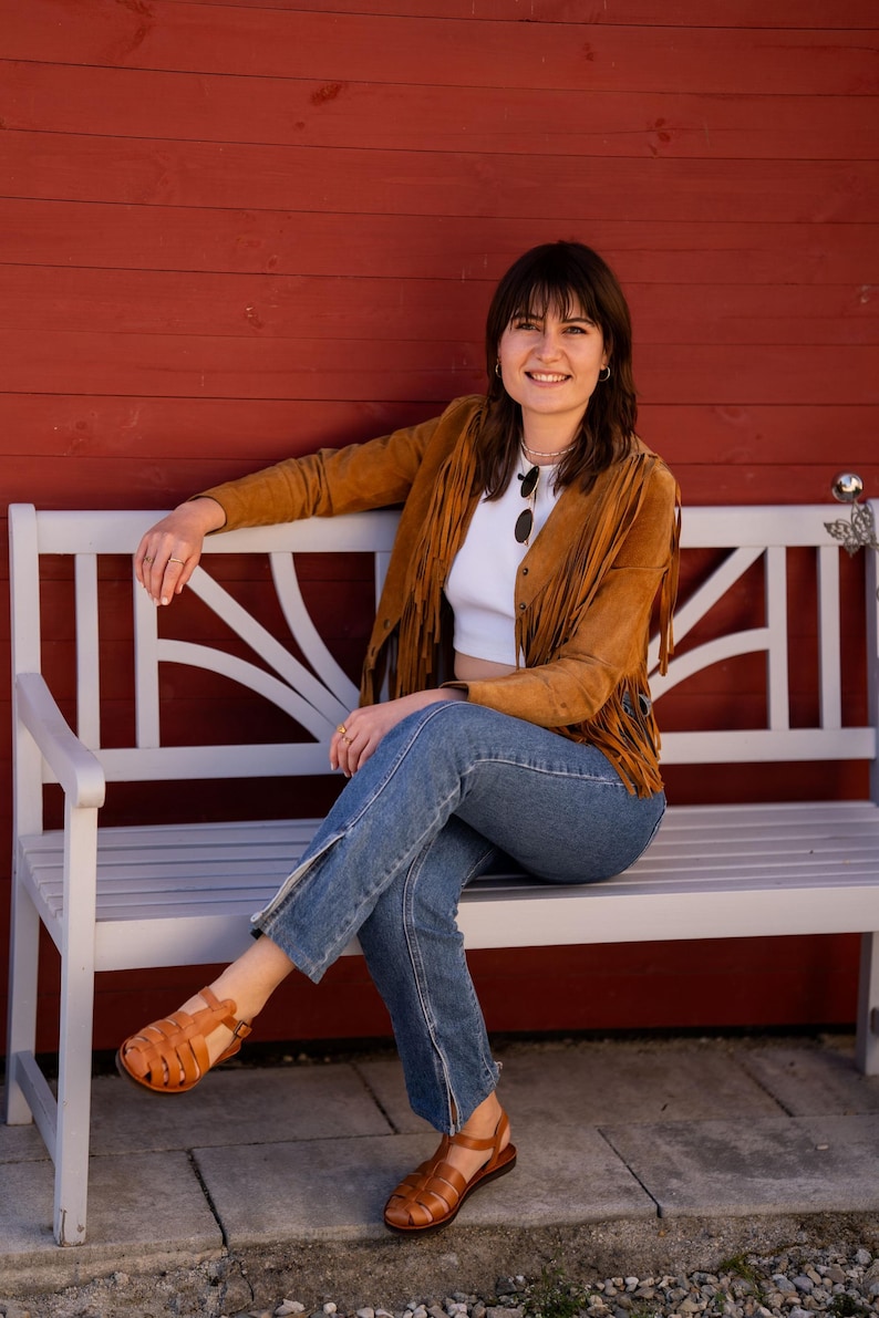 May include: A woman wearing a brown suede fringe jacket, white crop top, blue jeans, and brown sandals sits on a white wooden bench in front of a red wooden wall.