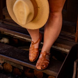 May include: A pair of brown leather sandals with a gladiator style design. The sandals are worn by a person standing on a set of wooden steps.