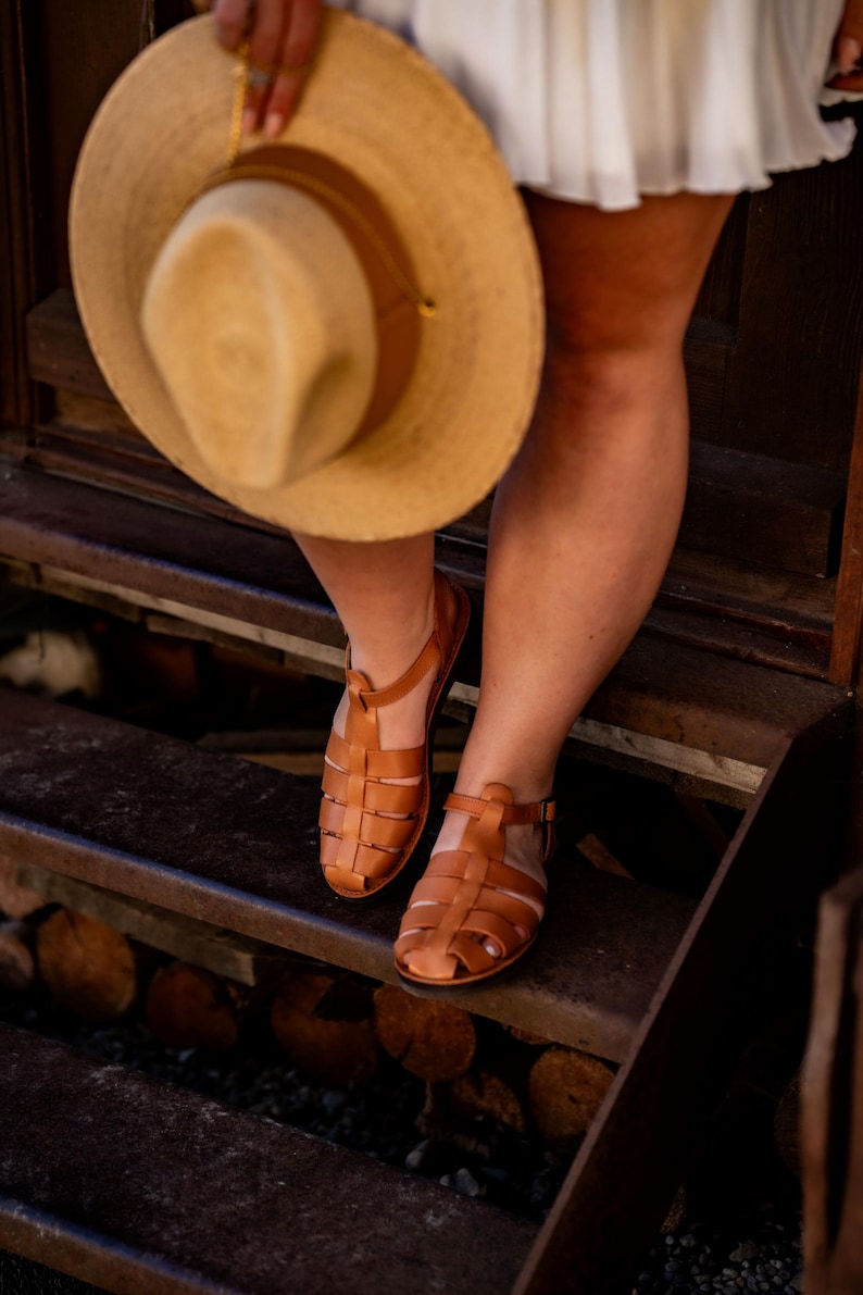 May include: A pair of brown leather sandals with a gladiator style design. The sandals are worn by a person standing on a set of wooden steps.