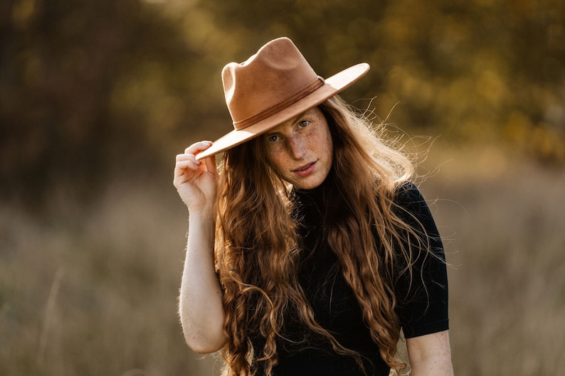 Peut inclure: Une femme aux longs cheveux auburn ondul&eacute;s porte un chapeau fedora brun. Elle porte un chemisier noir &agrave; col montant et se tient dans un champ d'herbe haute.