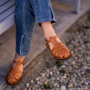 May include: A pair of brown leather sandals with a woven design. The sandals are worn by a person sitting on a bench.