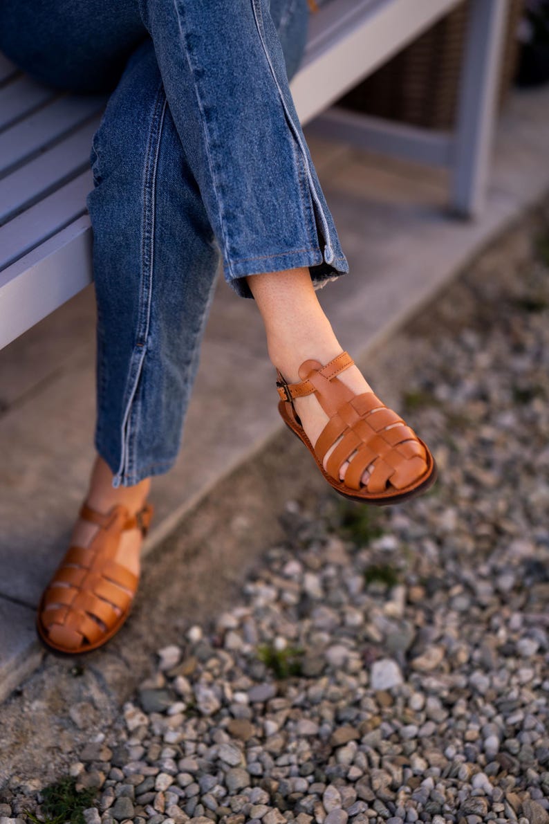 May include: A pair of brown leather sandals with a woven design. The sandals are worn by a person sitting on a bench.