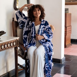 K&ouml;nnte beinhalten: Eine Frau in einem blau-wei&szlig; gemusterten Kimono mit wei&szlig;er Hose und braunen Sandalen sitzt auf einem Stuhl. Der Stuhl steht in einem Raum mit einer wei&szlig;en Wand und einem Spiegel.