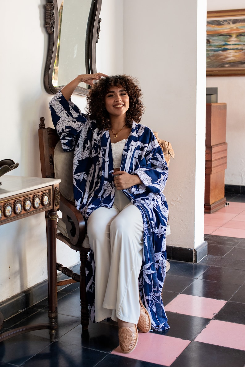 K&ouml;nnte beinhalten: Eine Frau in einem blau-wei&szlig; gemusterten Kimono mit wei&szlig;er Hose und braunen Sandalen sitzt auf einem Stuhl. Der Stuhl steht in einem Raum mit einer wei&szlig;en Wand und einem Spiegel.