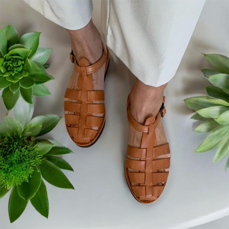 May include: Tan leather sandals with a woven design and an ankle strap. The sandals have a closed toe and a flat sole.
