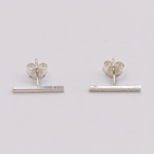 May include: A pair of minimalist silver bar stud earrings. Each earring features a straight, horizontal bar attached to a post with a butterfly clutch backing. The earrings are displayed on a white background.