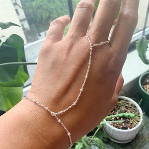 May include: A delicate silver hand chain bracelet. The bracelet connects from the ring finger to the wrist with a fine chain and small, round beads. The jewelry is displayed on a hand, with a blurred background of greenery and a window.