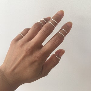 Pode incluir: Uma mão com vários anéis finos de prata nos dedos. Os anéis são espaçados uniformemente e parecem ser feitos de metal. Os dedos adornados com anéis incluem o indicador, o médio, o anelar, o mindinho e o polegar. Fundo branco liso.