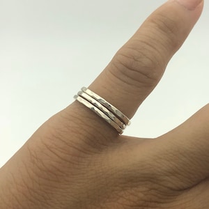 May include: Three silver hammered rings stacked on a finger. The rings have a textured surface and a simple, minimalist design. The rings are worn on a finger against a neutral background.