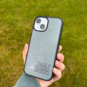 May include: A smartphone case with a gray topographic design and a black border. The case features the text "DAILY BREAD MUSIC" with a graphic of a turntable. The phone case is held against a background of green grass.