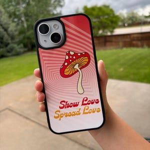 May include: A black iPhone case with a red and pink background featuring a mushroom illustration. The case has the words "Show Love Spread Love" in orange and yellow. The case is designed for an iPhone.