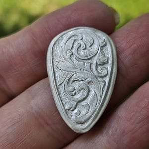 May include: A silver-colored, teardrop-shaped pendant with an intricate, engraved floral design. The pendant features a detailed pattern of swirling leaves and vines, showcasing a classic, elegant style. The edges are smooth and rounded.