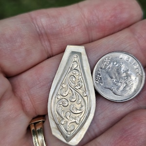 May include: A silver-colored, teardrop-shaped pendant with an ornate, swirling floral design. The pendant is next to a U.S. dime for size comparison. The pendant is held in a hand.