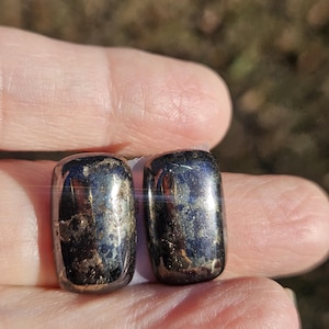 May include: Two polished, rectangular, dark metallic-colored stones. The stones have a reflective surface with areas of lighter, textured detail. The image is of a jewelry item, possibly earrings or pendants. The text "Littleredjewelrydesign" is visible at the bottom.